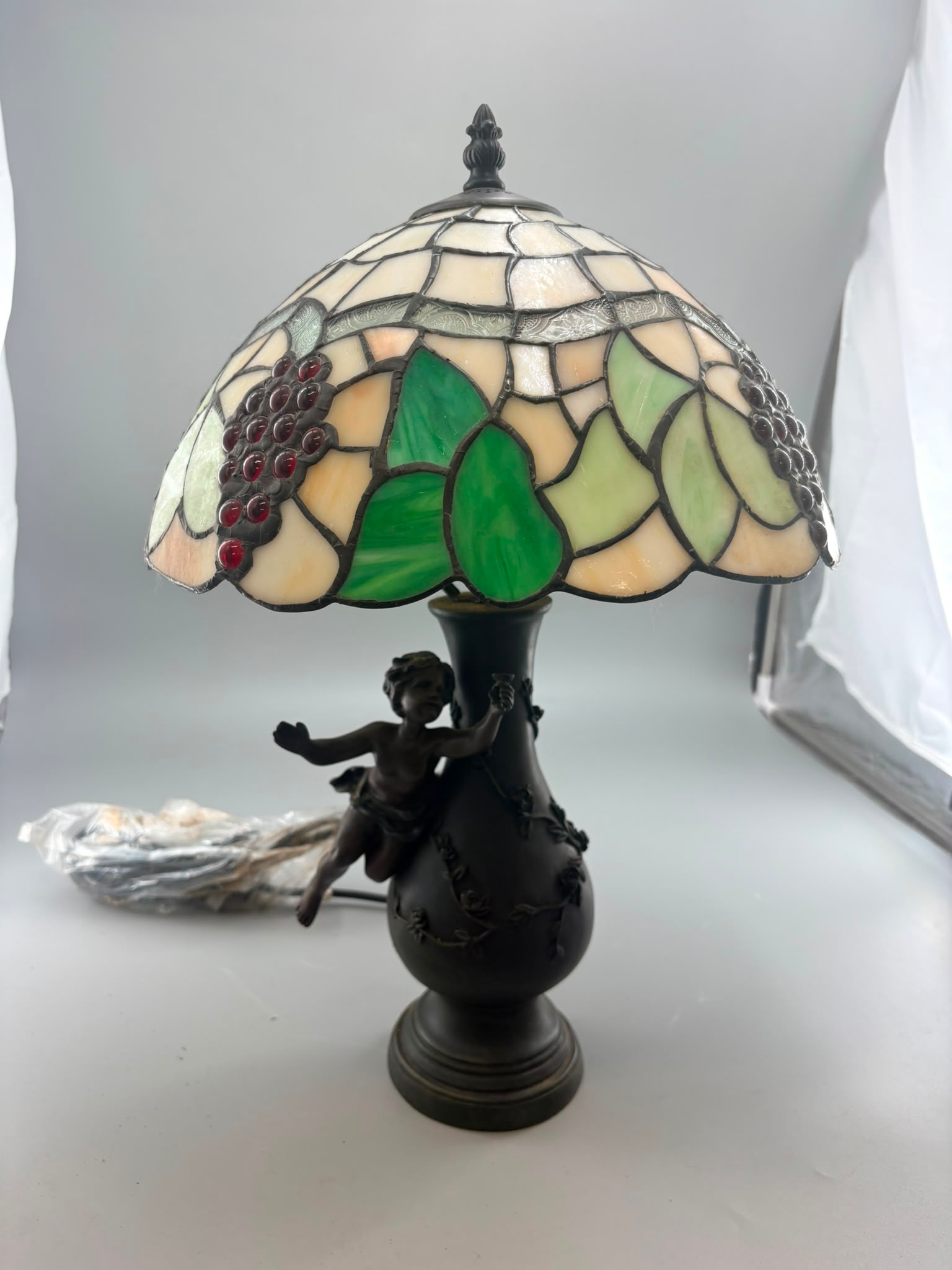 Tiffany-style table lamp with cream stained-glass shade, green leaf panels, dark red grape cabochons, and a dark cherub-mounted base